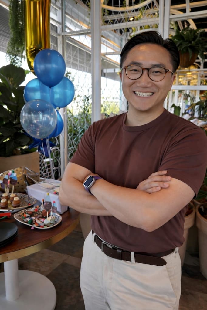Jason Lu smiling, wearing round glasses and a brown t-shirt, arms crossed.